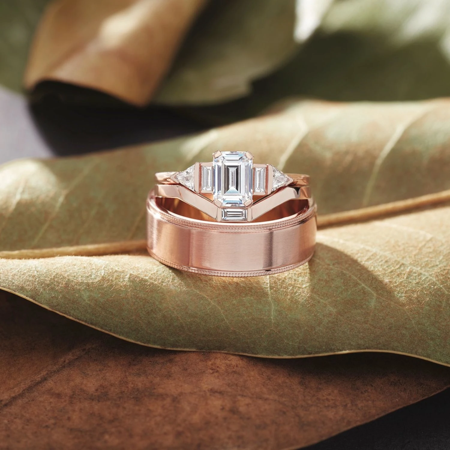 Rose gold emerald cut engagement ring with matching wedding band on green leaf